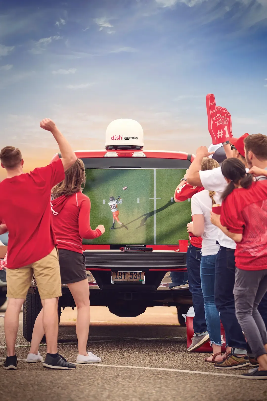 A group of fans cheer while watching a football game on a television mounted in the back of a red pickup truck equipped with a Dish Playmaker satellite.